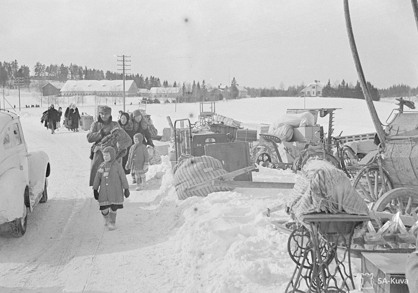 Perhe matkalla talvella 1944–46 — sotilas-isä kantaa kantamuksissaan vauvan, äiti vetää pikkulasta kädestä, etualla pieni tyttö katsoo suoraan kameraan. Mustavalkokuva SA-kuva-arkistosta.