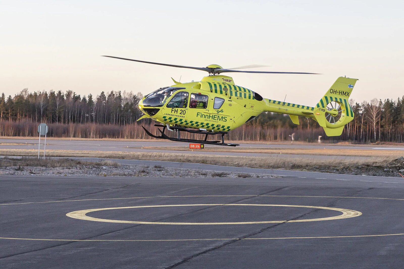 FinnHEMS-lääkärihelikopteri OH-HMX laskeutumassa tukikohdan kentälle hämärässä syysillassa. Keltavihreä Airbus EC135 -kone leijuu kentän yläpuolella, taustalla Lapin lehtimetsää ja vaaleansininen taivas.