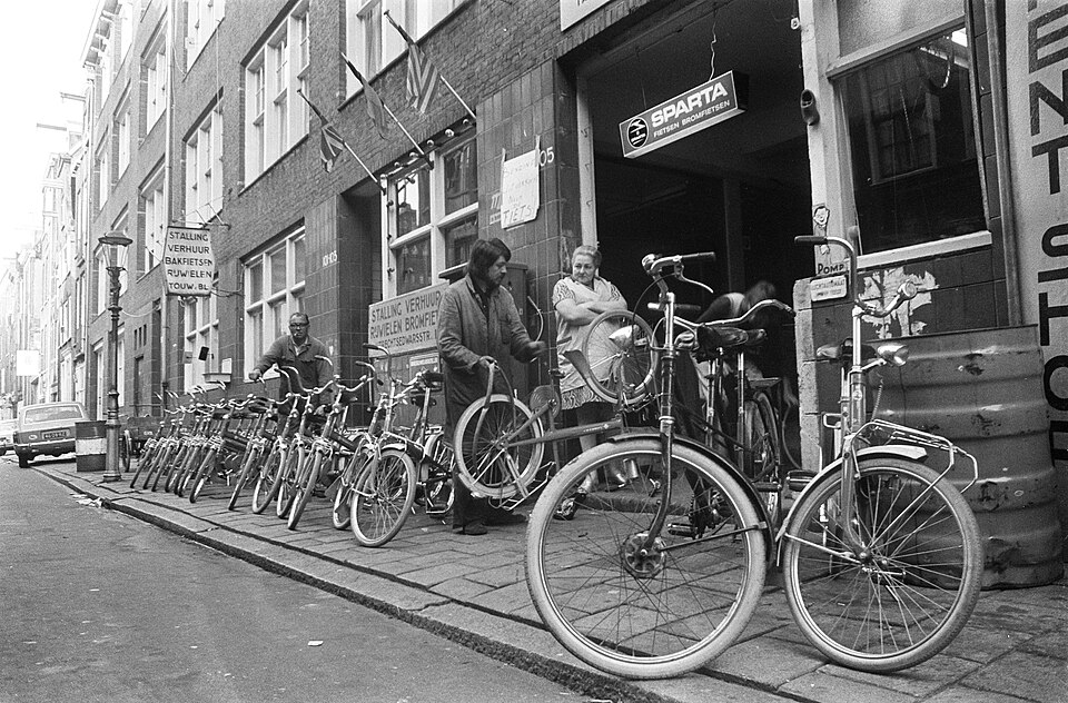 Pyörävuokraamo Amsterdamissa marraskuussa 1973. Mustavalkoinen valokuva: rivi polkupyöriä jalkakäytävällä, vuokraajat ja pari asiakasta liikkeen edessä, sisäänkäynnin yllä Sparta-fietsen-kyltti.